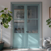 A pair of internal doors with clear glass panels, primed and painted in a sage sky color, installed in a room with a gray floor and walls, alongside a potted plant and a chair.