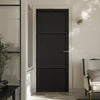 A prefinished black internal door with three panels, installed in an interior setting with light walls and a visible light fixture above the door.